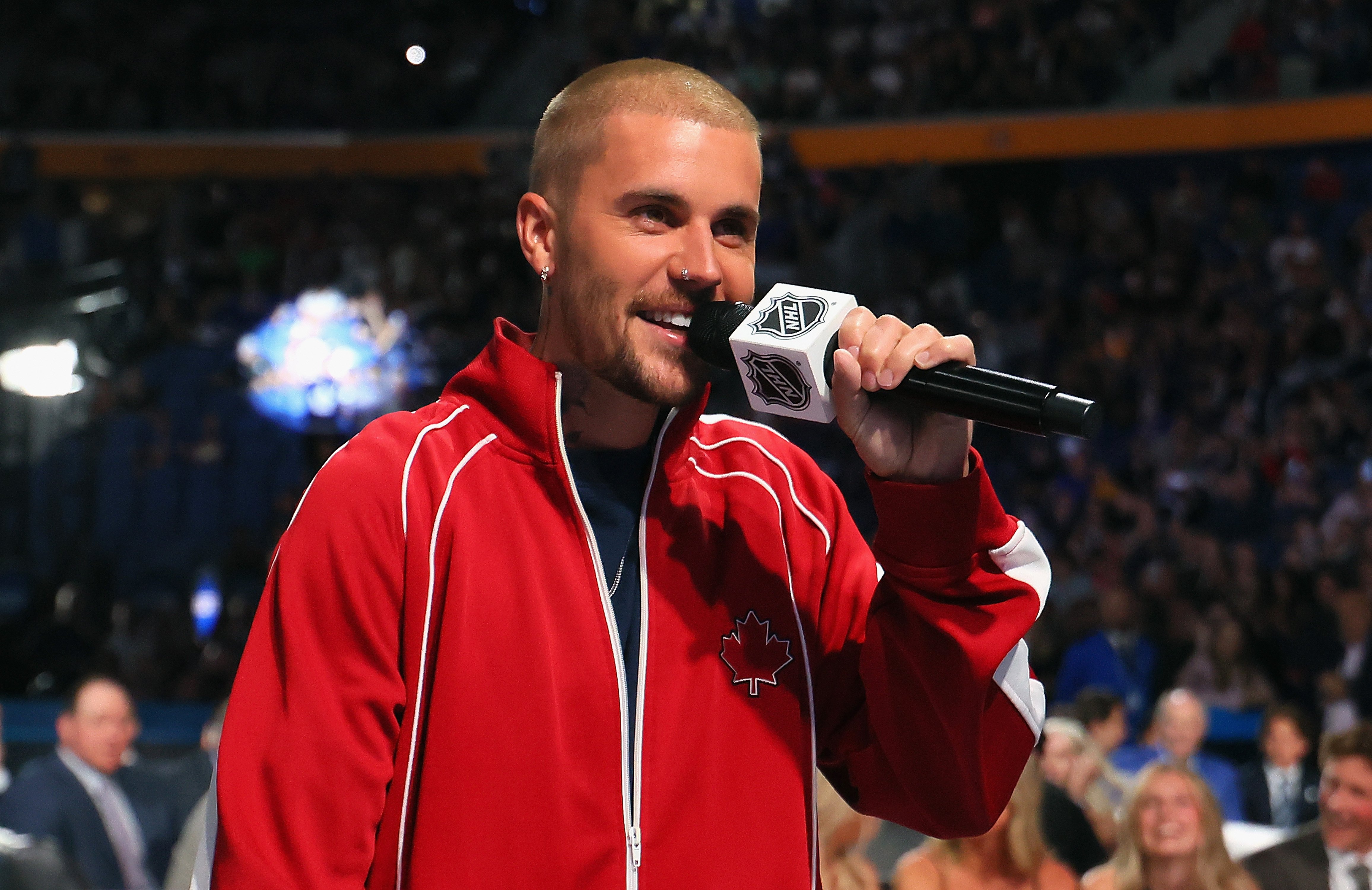 A man with a shaved head and a red tracksuit featuring a maple leaf logo speaks into an NHL-branded microphone in front of a crowd.