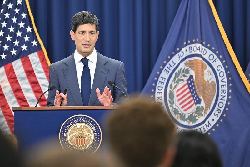 A man in a dark suit and tie speaks at a podium bearing the Federal Reserve Board of Governors seal, flanked by an American flag and a Federal Reserve flag.