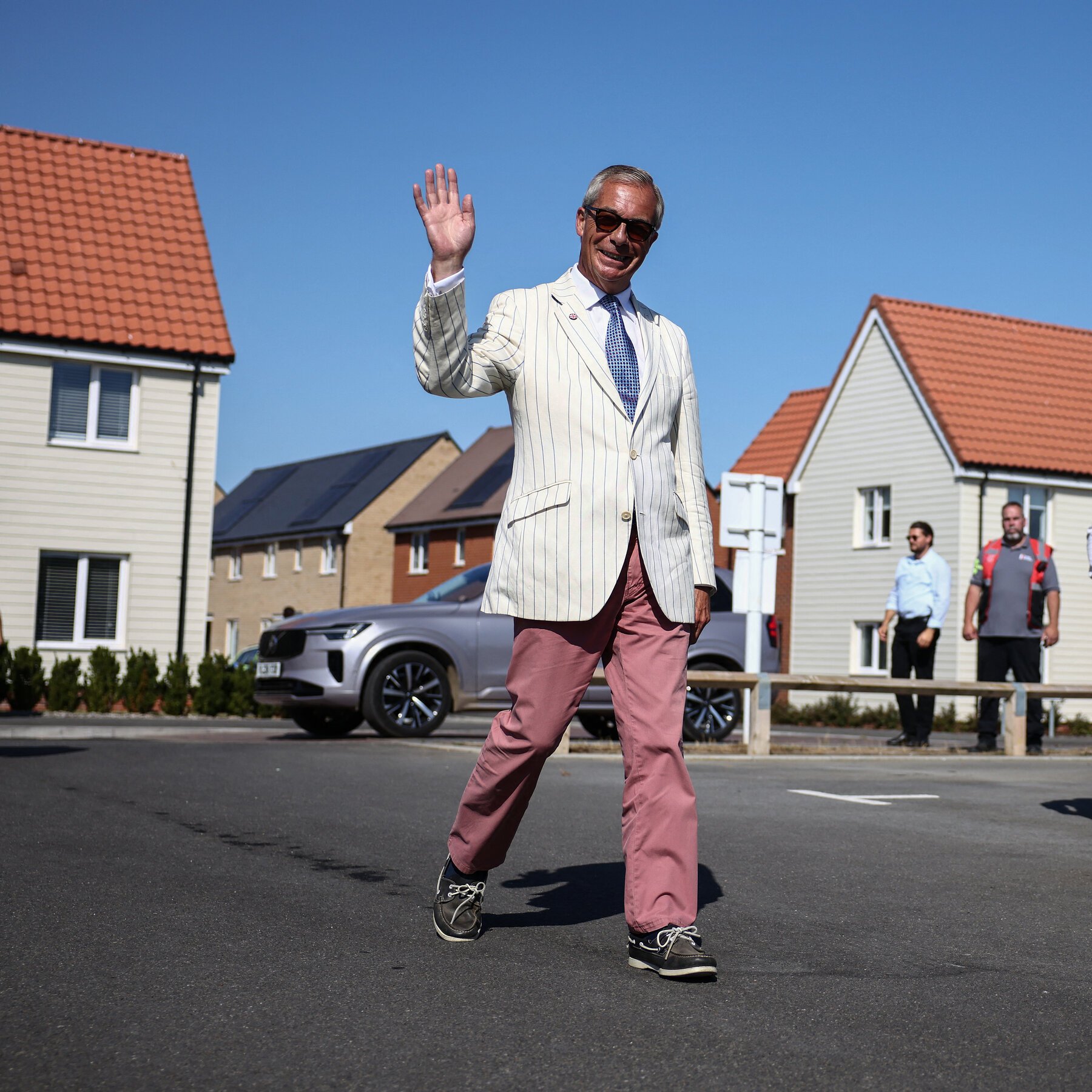 A man in a white pinstripe blazer, pink trousers, and sunglasses waves while walking down a residential street lined with houses and a parked SUV.