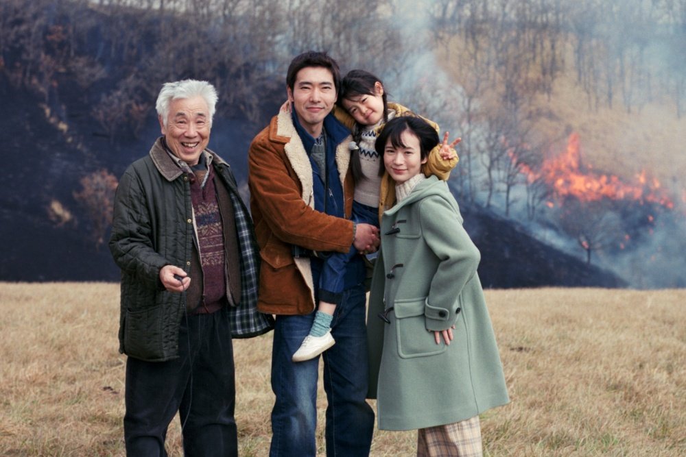 Four people smile and pose in a grassy field with smoke and flames rising from a wooded hillside in the background.