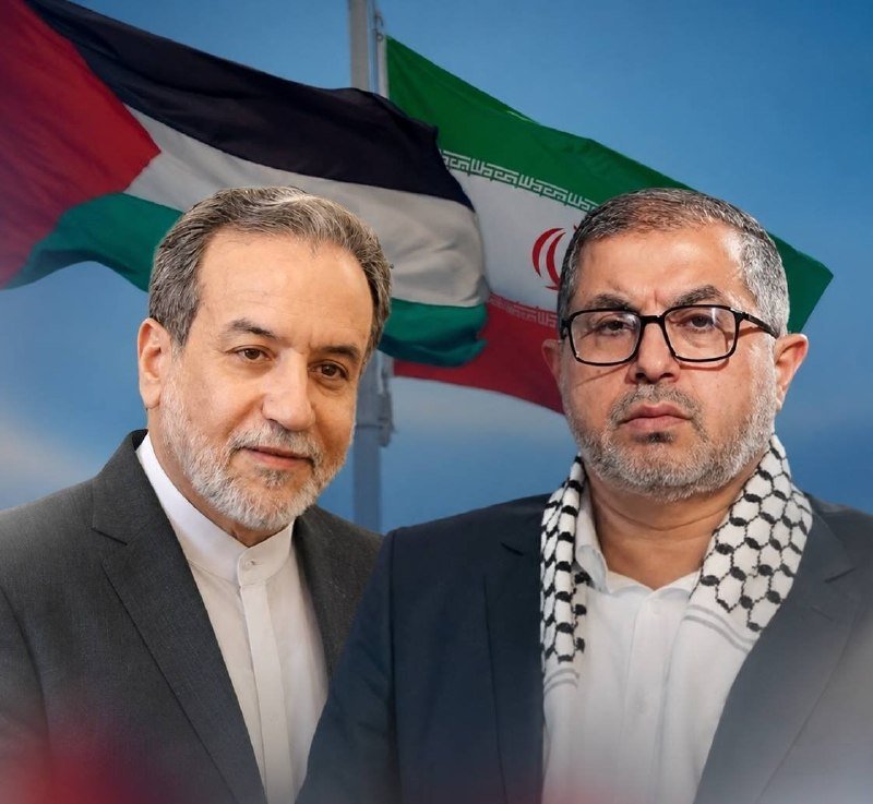 Two men pose in front of Palestinian and Iranian flags against a blue sky backdrop.