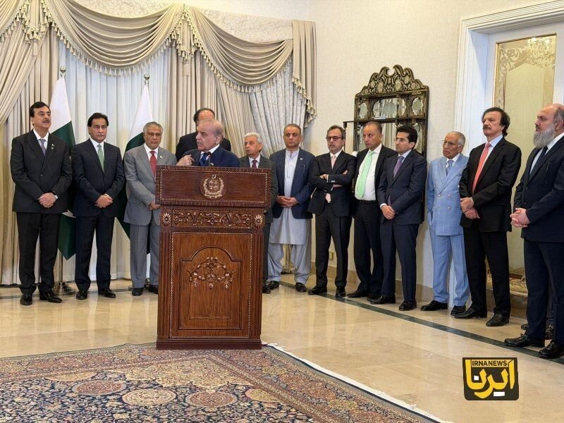 A man speaks at an ornate wooden podium bearing an emblem while eleven men in formal attire stand behind him in a room with draped curtains and a Pakistani flag.