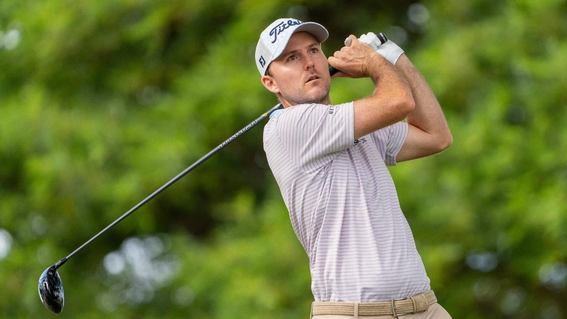 A golfer in a white Titleist cap and striped shirt follows through on a swing against a green foliage backdrop.