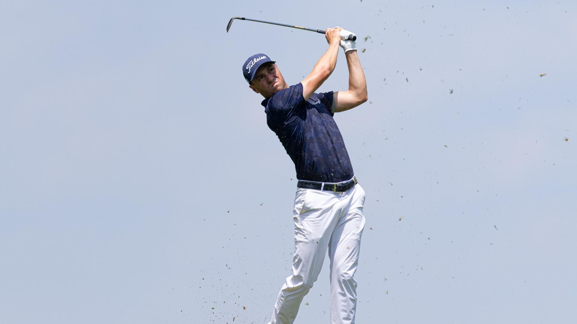A golfer in a blue "Lineage" polo and black "Titleist" cap watches a shot while holding a wedge, with blurred spectators visible behind him.