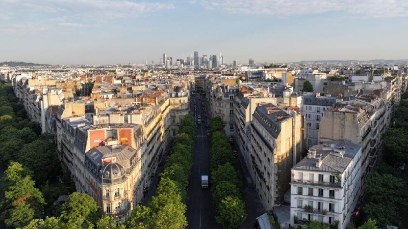 Aerial view of a wide tree-lined boulevard cutting through dense Parisian-style buildings, with a distant cluster of modern skyscrapers visible on the horizon.