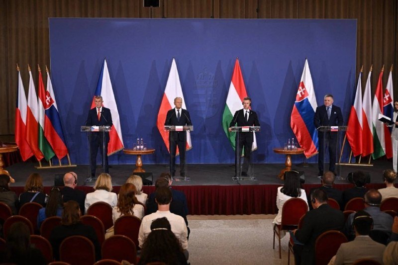 Four men in suits stand at podiums on a stage backed by national flags and a coat of arms, addressing a seated audience.