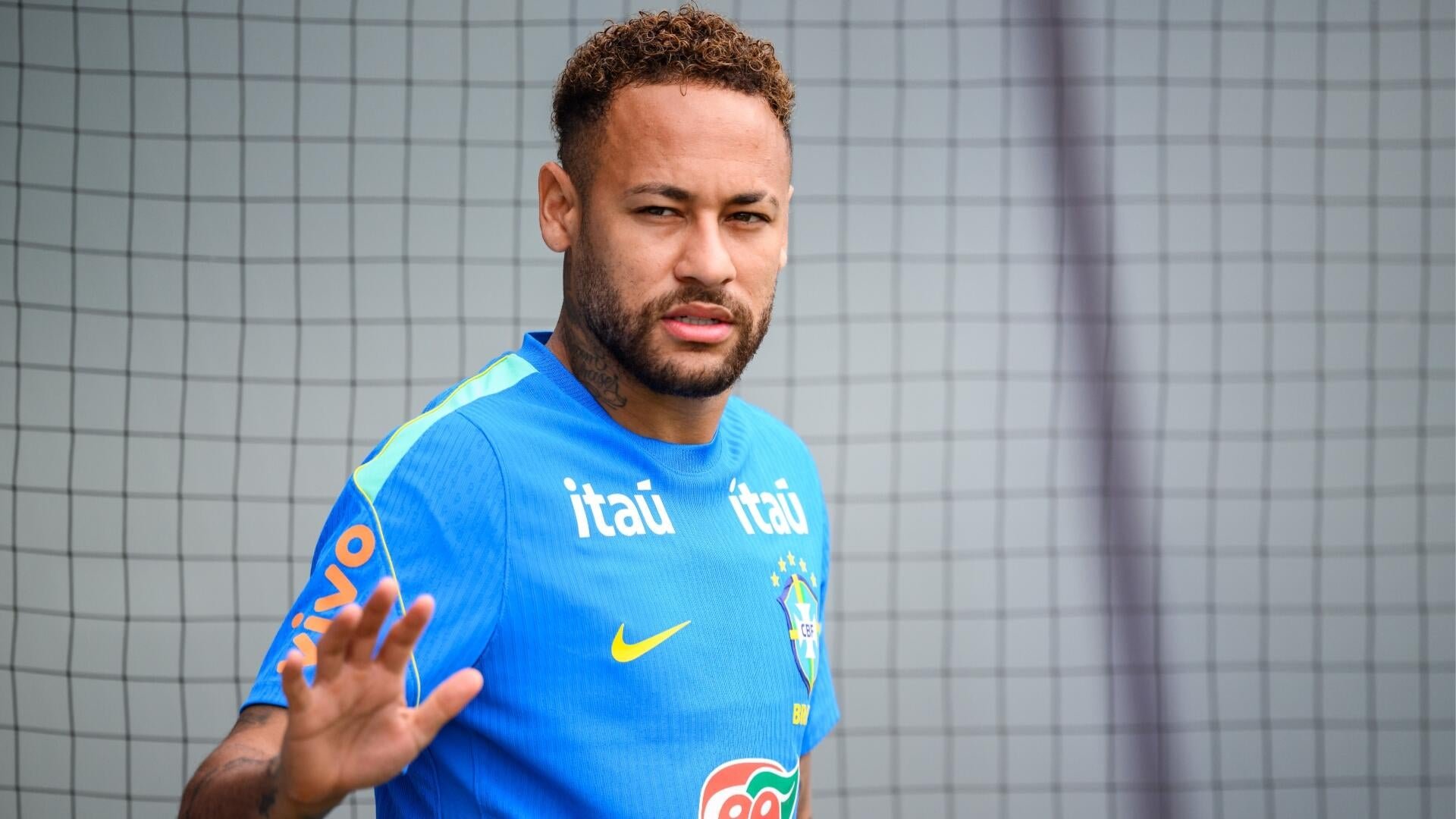 A man with curly hair and a beard, wearing a blue Brazil national team training jersey with "Itaú" sponsorship, stands indoors near a netted structure.