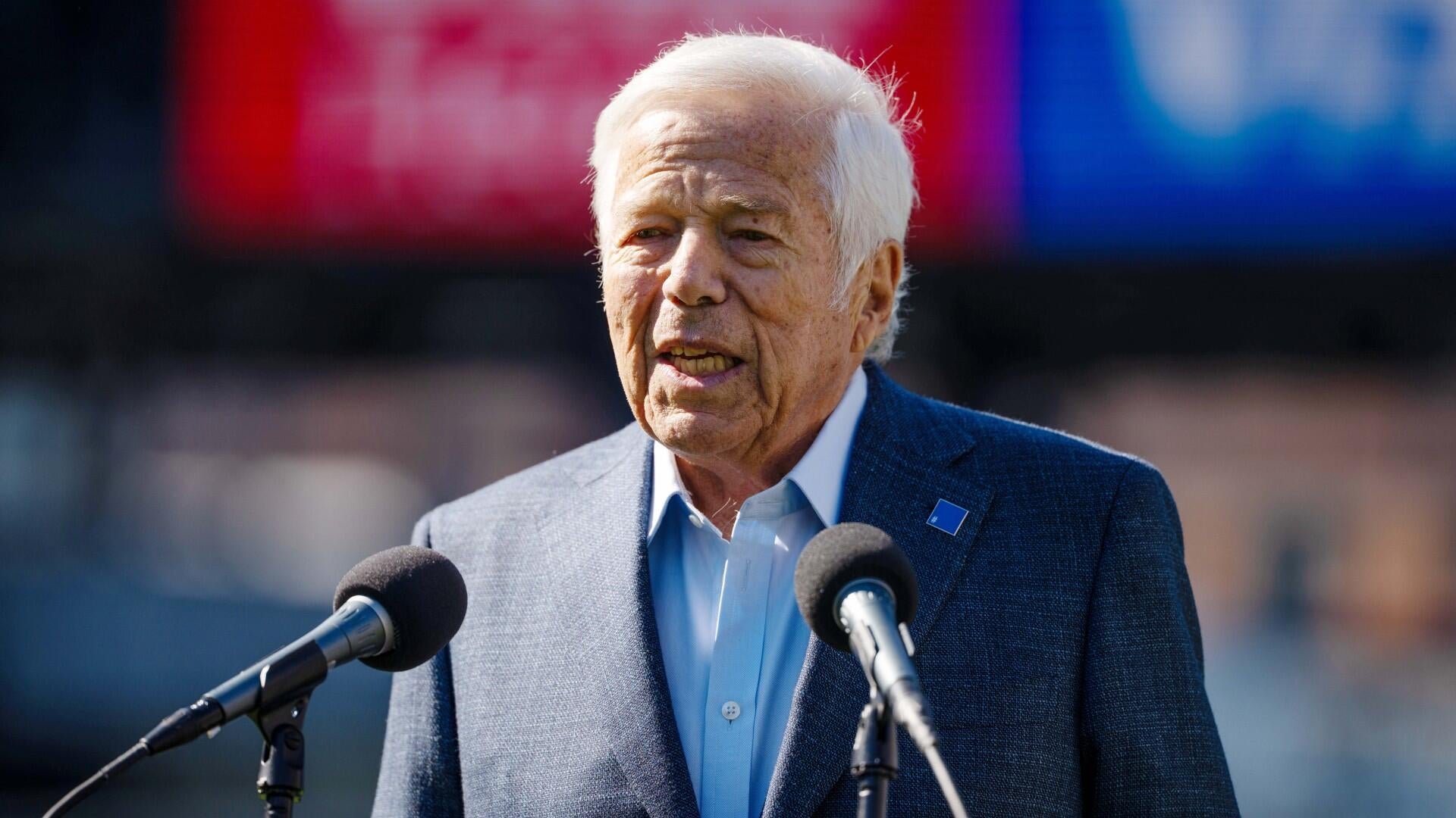 An elderly man with white hair speaks into two microphones, wearing a dark blazer and light blue shirt at an outdoor event.