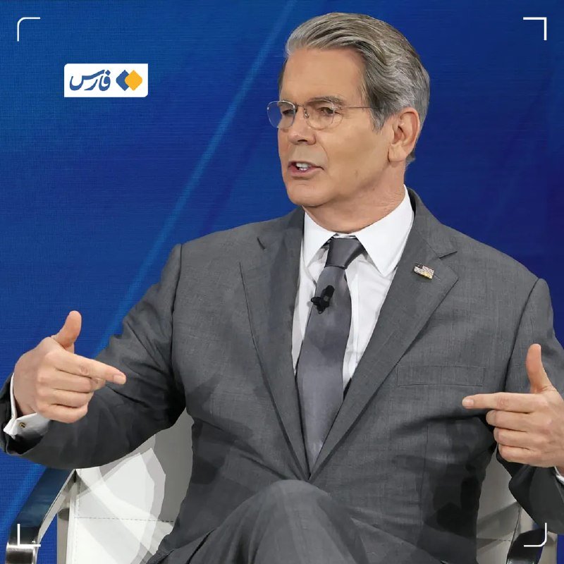 A gray-haired man in glasses, a gray suit, and an American flag pin speaks while gesturing with both hands against a blue backdrop.