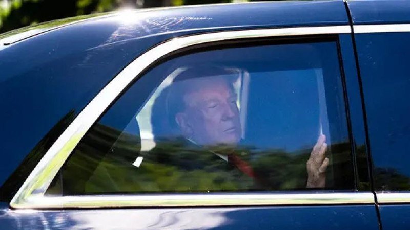 A man in a suit and red tie sits inside a dark vehicle, visible through a tinted window, with his hand raised near the glass.