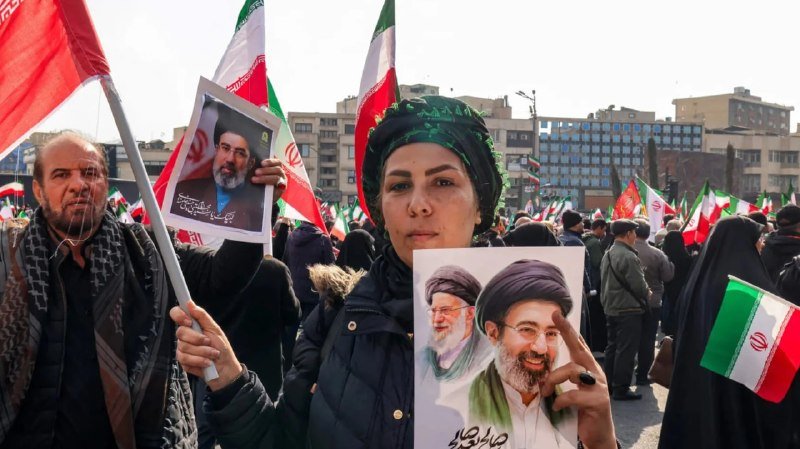 A crowd gathers holding Iranian flags and posters featuring portraits of Iranian officials, with a woman in a black headscarf making a victory sign in the foreground.