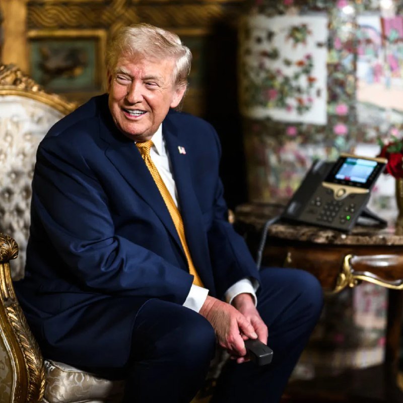 A smiling older man with light hair, wearing a dark navy suit and gold tie, sits in an ornate gilded chair beside a desk phone in a lavishly decorated room.
