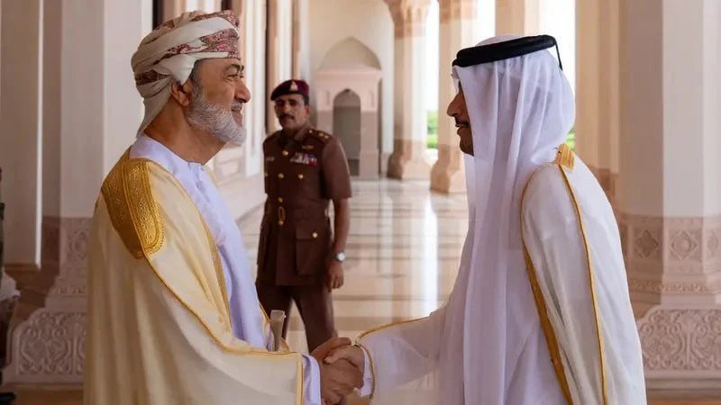 Two men in traditional Arab attire shake hands and smile in a columned hall, while a uniformed officer stands in the background.