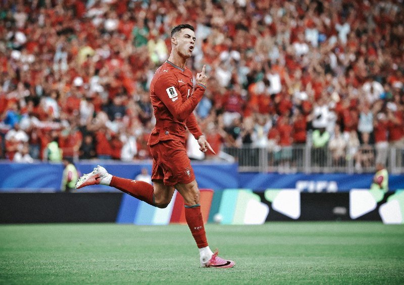 A soccer player in a red uniform runs across a grass field, pointing upward with one finger in celebration before a packed stadium crowd.