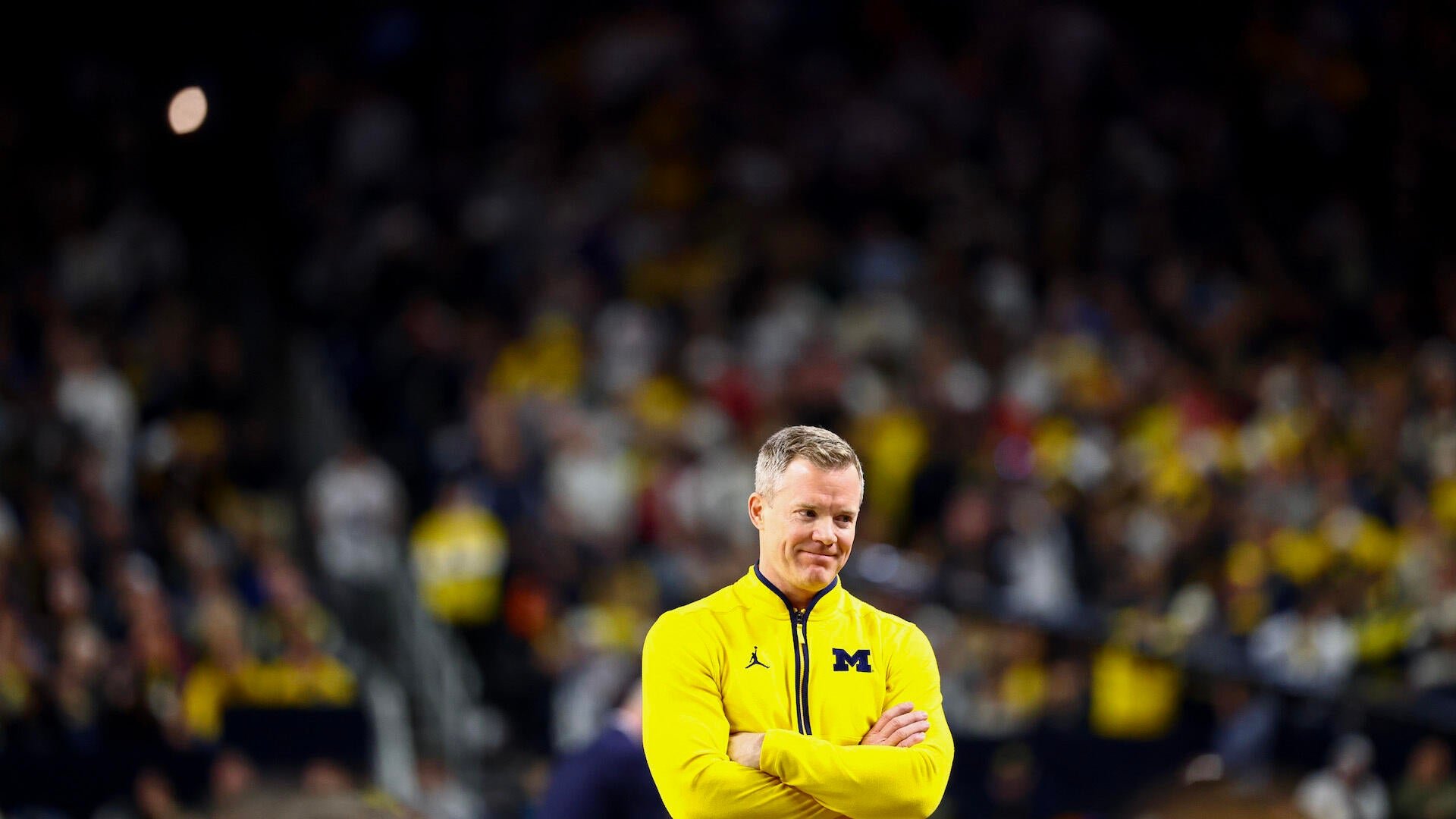 A man in a yellow quarter-zip pullover with a "M" logo stands with crossed arms in front of a blurred arena crowd.