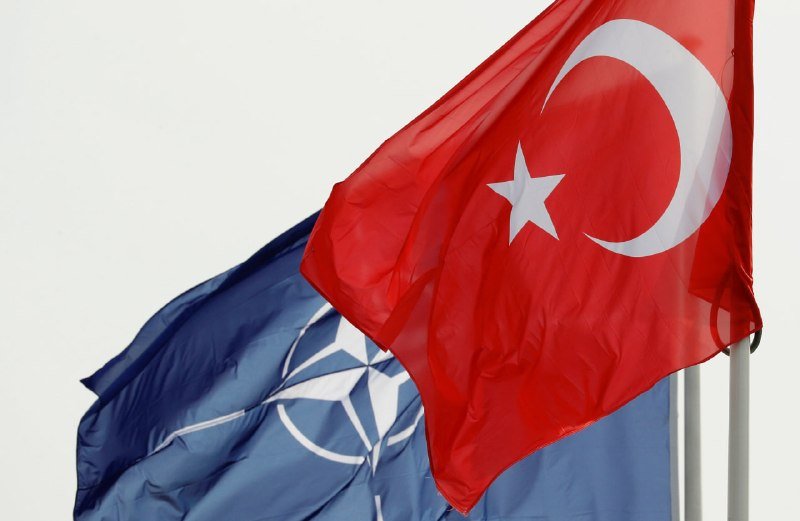 Two flags wave against a light sky: a blue NATO flag with its white compass emblem, partially obscured by a red Turkish flag bearing a white crescent and star.