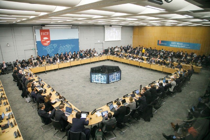Numerous attendees sit around a large square conference table with laptops, facing a central video display in a room displaying "FATF" signage.