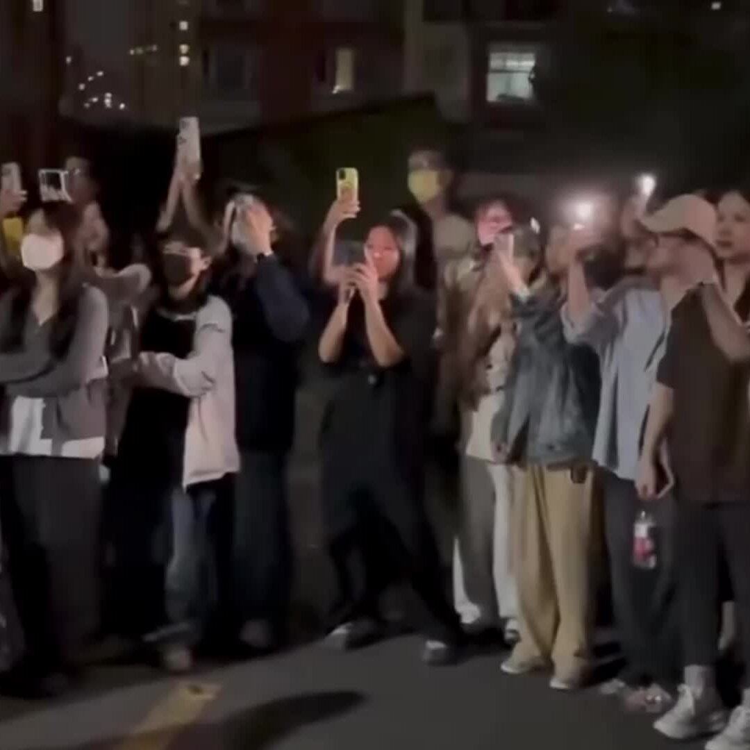 A crowd of masked people at night hold up smartphones with flashlights on, appearing to record or photograph something out of frame.