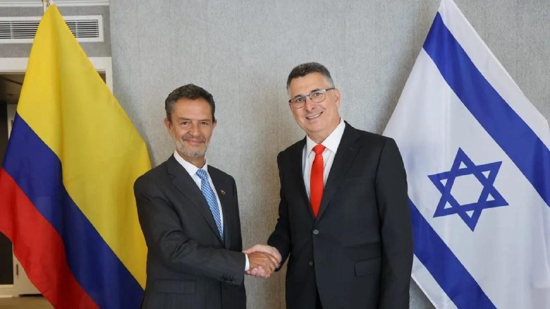 Two men in suits shake hands in front of the Colombian and Israeli flags.