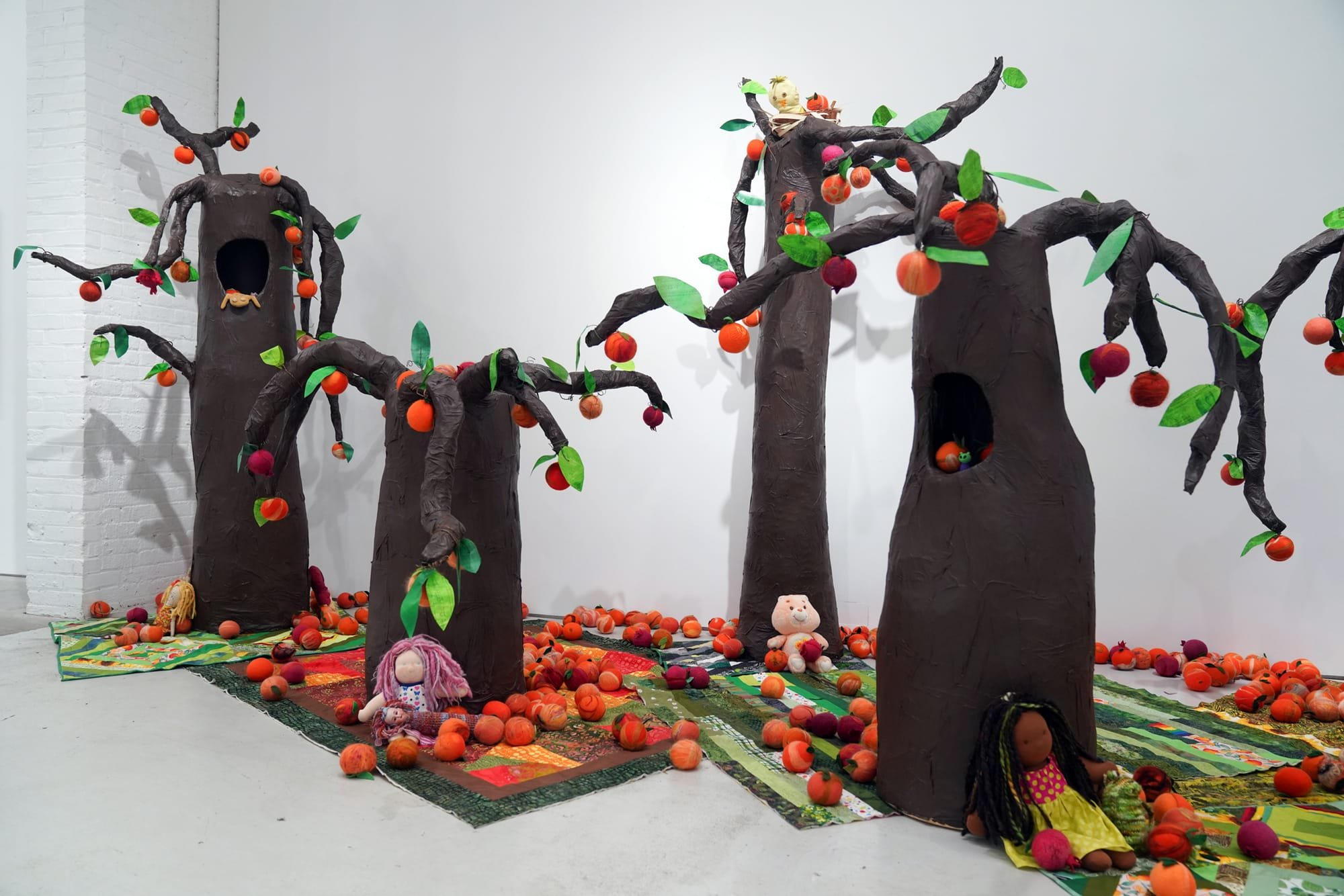 An art installation features brown sculptural trees with colorful fabric fruits, leaves, and toy figures arranged on patterned quilts covering the gallery floor.
