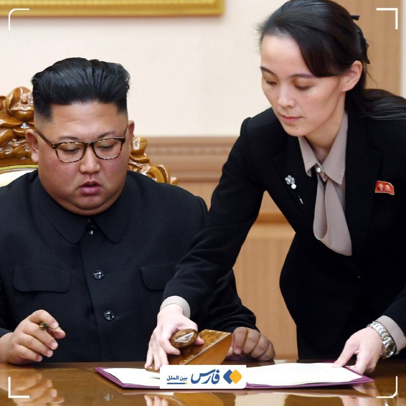 A man with glasses in a dark suit sits at a wooden desk while a woman in a dark blazer leans over to point at documents on a purple folder.