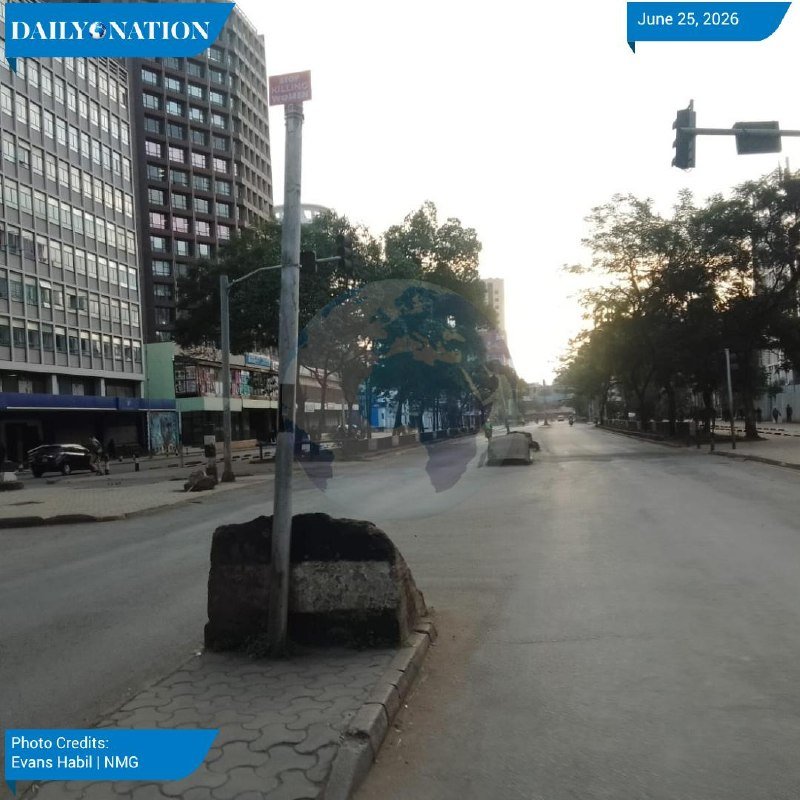 An empty city street at dawn with a traffic light, traffic signal pole, concrete median barrier, and multi-story buildings under a bright sky.