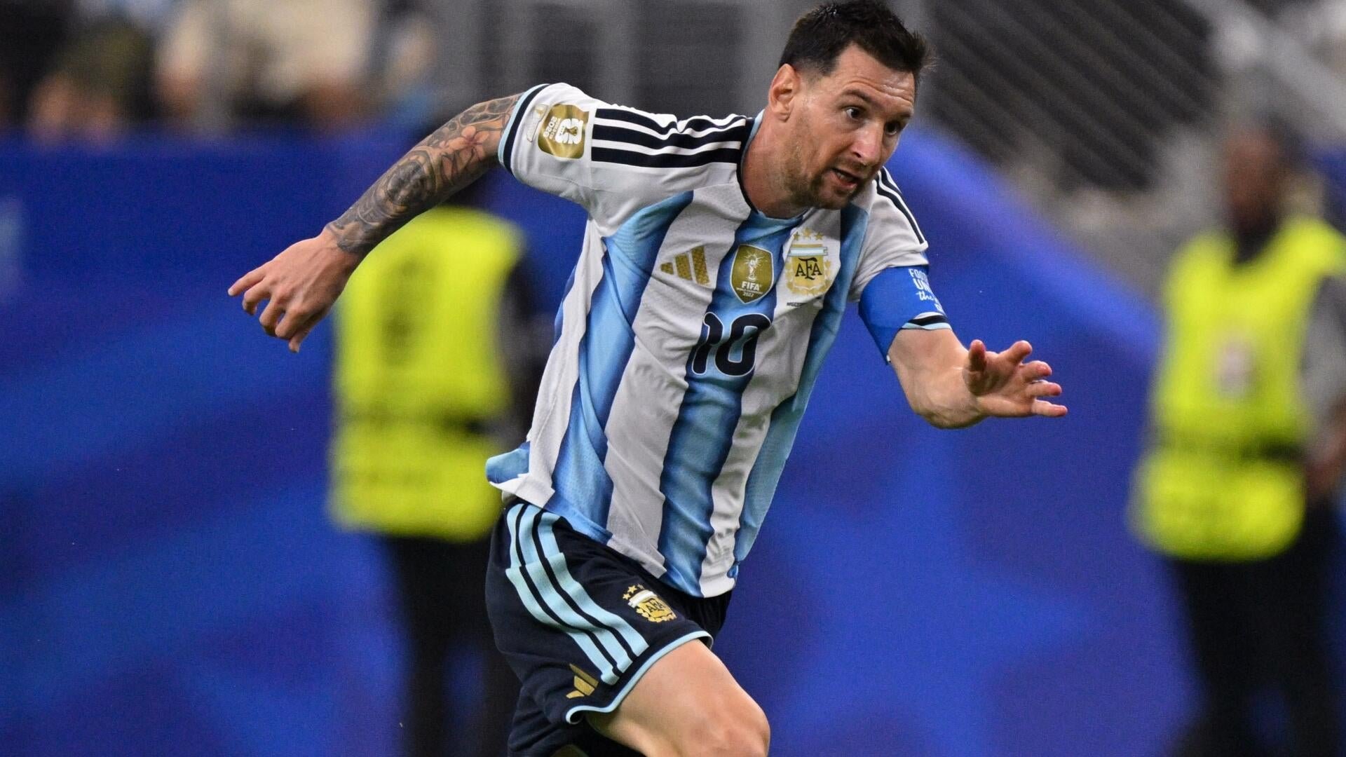 A tattooed soccer player wearing a blue-and-white striped Argentina jersey with the number 10 runs during a match, with blurred figures in yellow vests behind him.
