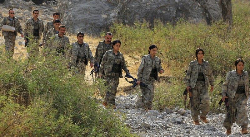A woman carrying a yellow flag walks past a large outdoor mural depicting several bearded men in black turbans and military attire.