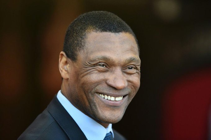 A man in a dark suit, white shirt, and blue tie smiles broadly against a blurred background.