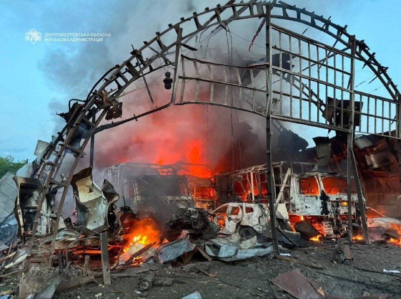 A large arched metal structure is engulfed in flames and thick smoke, with burnt debris and a damaged vehicle scattered across the ground below.