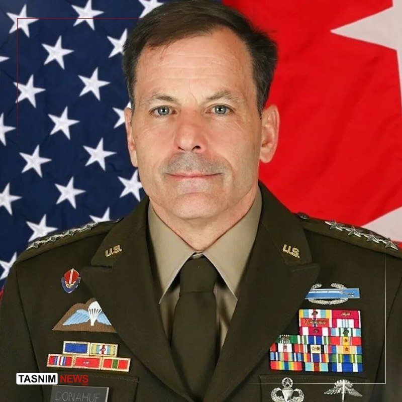 A smiling man in a dark suit and red tie stands with both arms raised, giving two thumbs up beside an American flag at a campaign-style event.