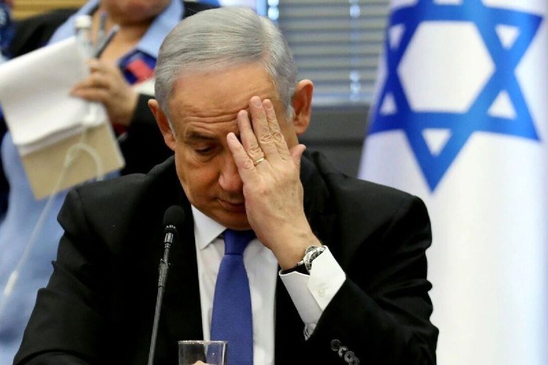 A gray-haired man in a dark suit and blue tie rests his hand against his forehead while seated at a table with a microphone, an Israeli flag visible behind him.