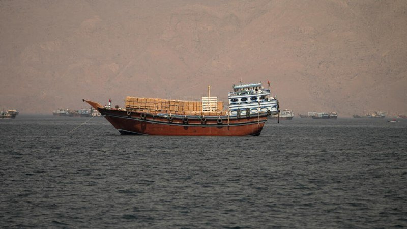 A large wooden cargo boat loaded with stacked boxes floats on gray water, with several smaller vessels visible in the distance against a backdrop of hazy mountains.