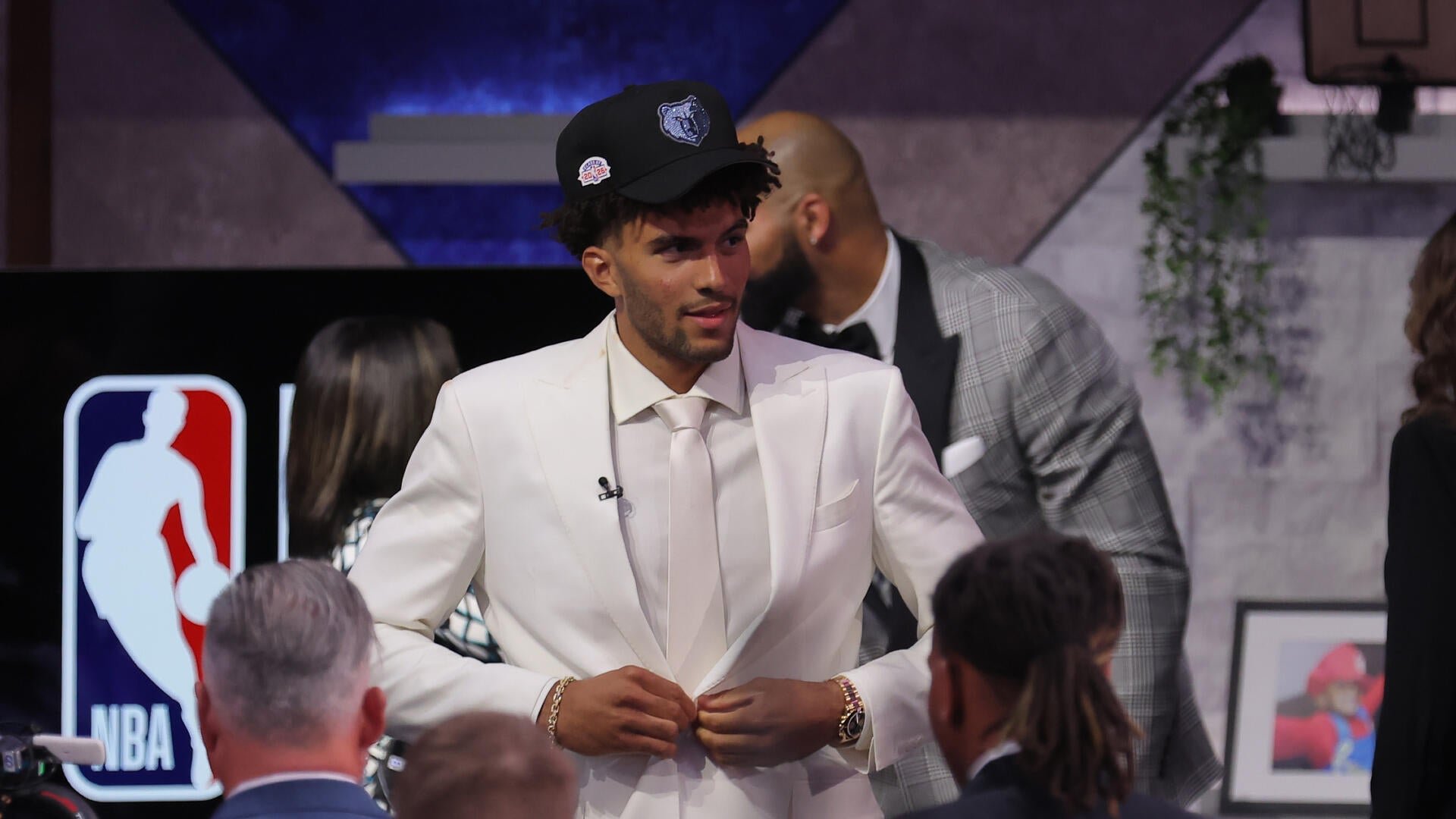 A large group of formally dressed young men and women pose together on stage in front of a brightly lit "NBA Draft 2026" logo, with a basketball held at center.