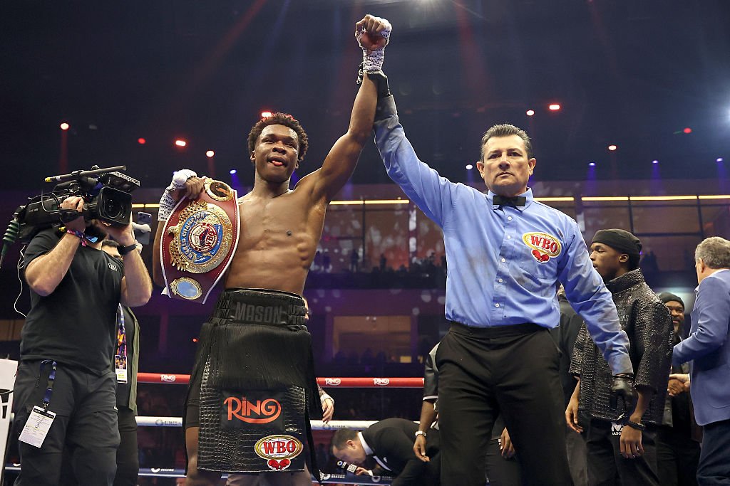 A shirtless boxer wearing black trunks labeled "MASON" holds a championship belt while a referee in a blue shirt raises his arm in victory inside a boxing ring.