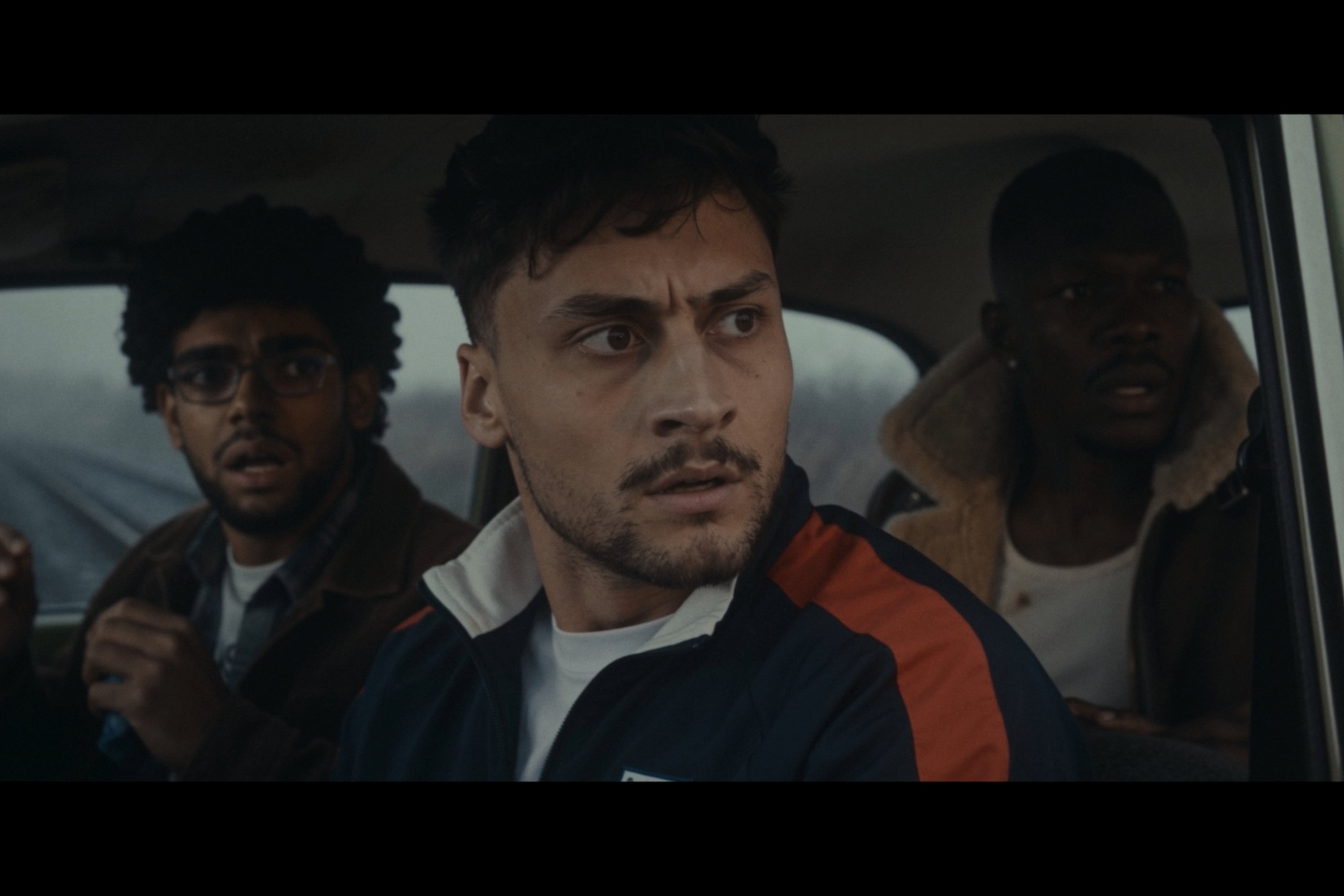 Three men sit inside a vehicle, with the central figure in a navy and orange jacket staring intently forward while two others look on with serious expressions.