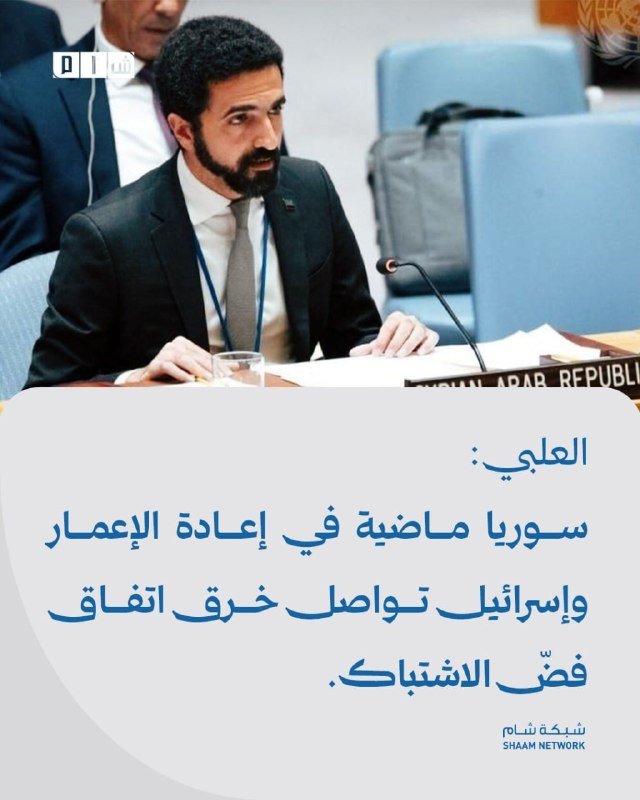A bearded man in a dark suit speaks at a microphone during a meeting, with a Syrian Arab Republic placard visible, accompanied by Arabic text and a "Shaam Network" logo.