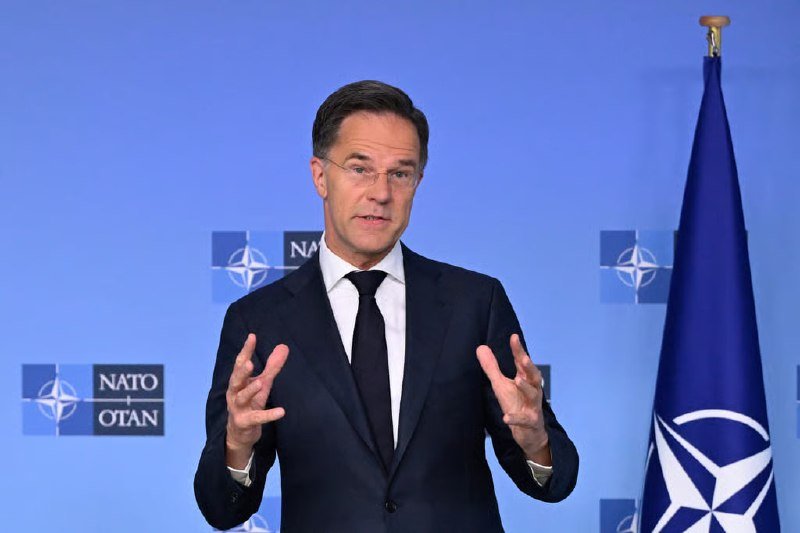 A man in a dark suit and glasses gestures with both hands while speaking in front of a blue backdrop displaying NATO logos and a flag.