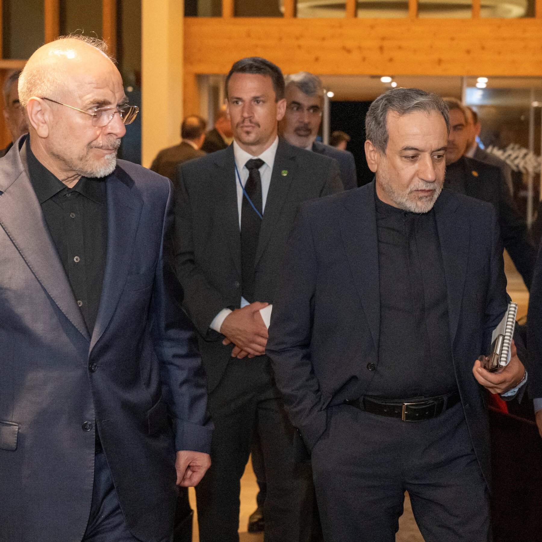 Two older men in dark suits walk together through a doorway, followed by other individuals in formal attire.