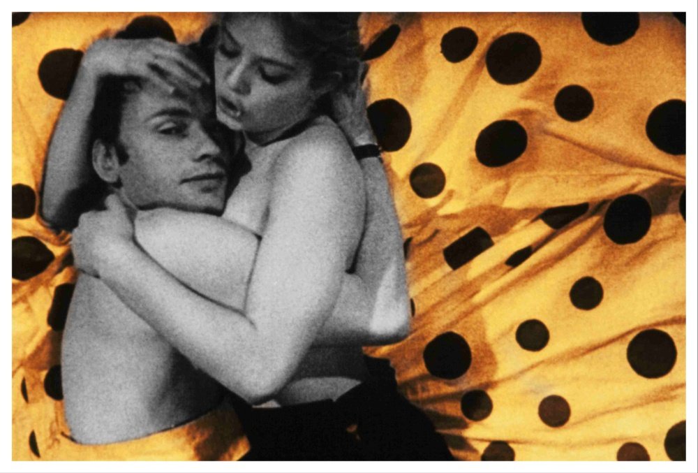 A shirtless man and a woman embrace against a black-and-white photograph layered over a polka-dotted orange fabric background.
