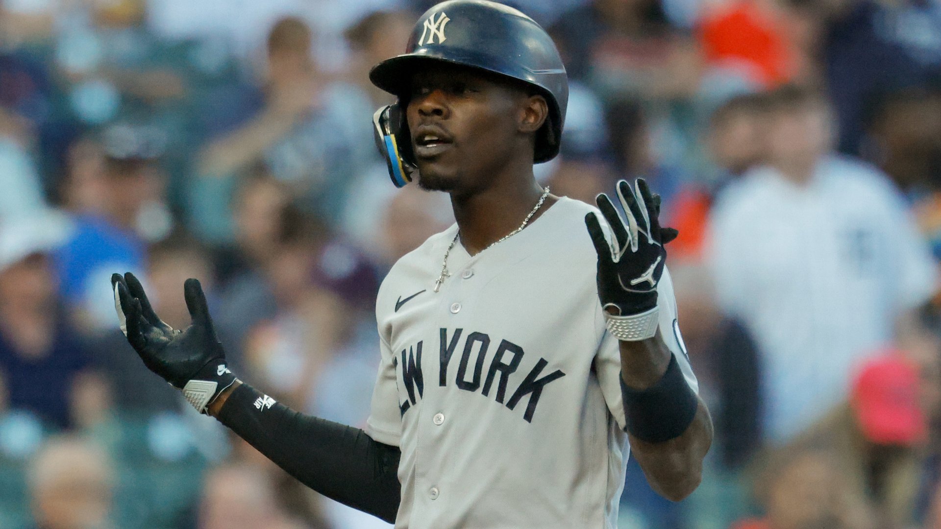 A baseball player in a New York Yankees uniform and navy batting helmet stands with both gloved hands raised, a blurred crowd visible behind him.