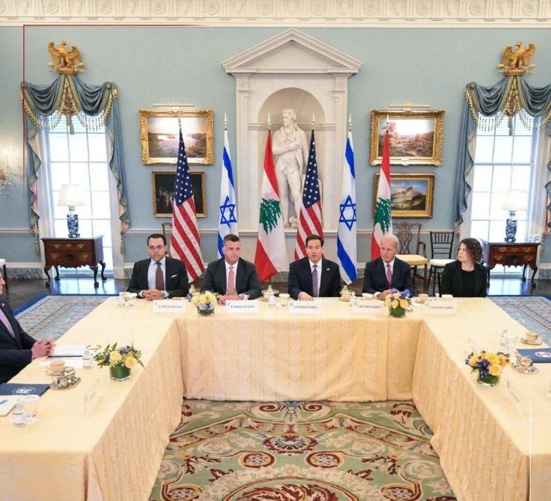 Five officials in business attire sit at a U-shaped table arrangement in an ornate blue room, with American, Israeli, and Lebanese flags displayed behind them.