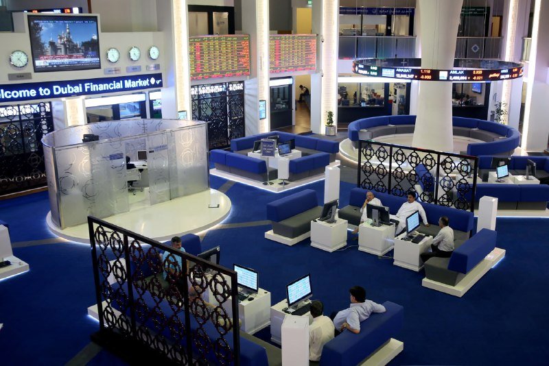 Several people work at computer stations on a blue-carpeted trading floor beneath digital boards displaying stock prices and a "Dubai Financial Market" sign.