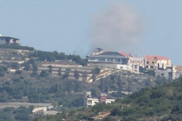 Dark smoke rises from a building on a rural hillside surrounded by scattered houses, trees, and dry terrain under a hazy sky.