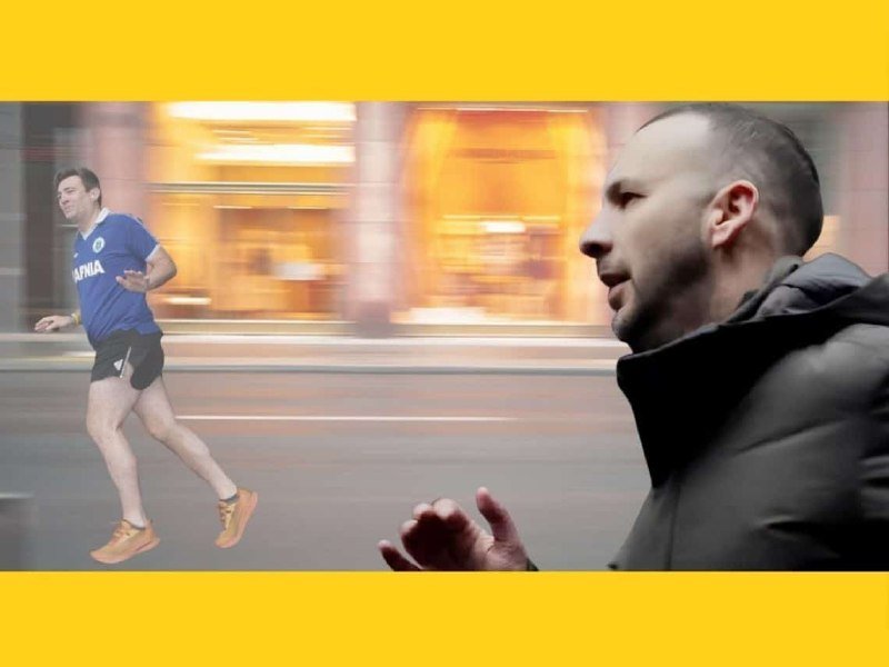 Two men run along a street, one in a blue shirt captured mid-stride in the background while another in a black jacket appears in close-up foreground, with a motion-blurred storefront behind them.