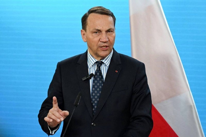 A man in a dark suit and patterned tie speaks at a podium, gesturing with his right hand, with a red and white flag visible behind him against a blue backdrop.