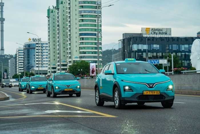 A line of turquoise VinFast SUVs with illuminated "TAXI" signs drives along a wet city street, passing an "Alatau City Bank" building and high-rise structures.