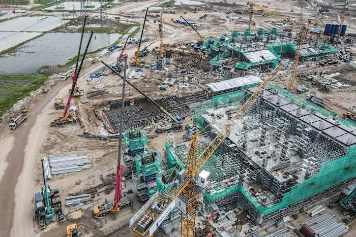 An aerial view shows a large active construction site with multiple cranes, scaffolding, and partially built structures covered in green netting, surrounded by dirt terrain.
