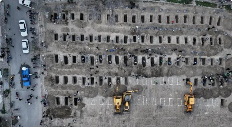 Aerial view of a cemetery with rows of freshly dug rectangular graves and excavators working in an adjacent paved lot.