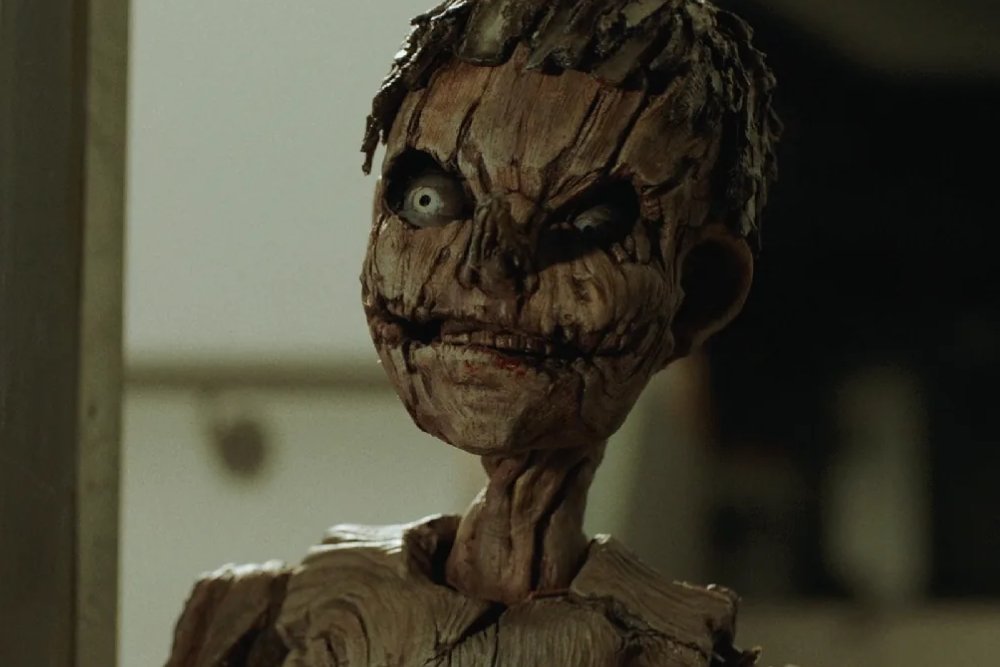 A close-up shows a small, humanoid figure made of textured, bark-like material with wide pale eyes and a stitched mouth, standing indoors.